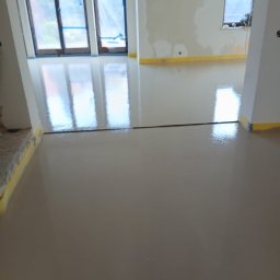 Świeżo wylana, gładka posadzka w jasnym odcieniu szarości, z widocznymi refleksami światła. Żółta taśma zabezpieczająca przy ścianach i oknach. W tle okna zakryte folią.