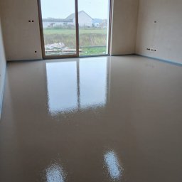 Gładka, świeżo wylana posadzka w jasnym pomieszczeniu z dużym oknem przesuwnym. Widoczne zabezpieczenia przy ścianach. Powierzchnia odbija światło.