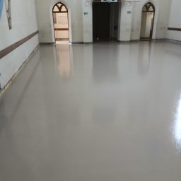 Gładka, jasna posadzka w przestronnym wnętrzu z łukowymi przejściami. Widoczne detale architektoniczne i zabezpieczenia przy ścianach. Nowoczesna wylewka w zabytkowym budynku.