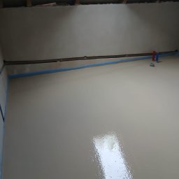 Świeżo wylana, gładka posadzka w jasnym odcieniu szarości, z widocznymi instalacjami grzewczymi wzdłuż ścian. Światło odbija się od mokrej powierzchni.