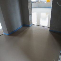 A&M Mariusz Batycki - Świeżo wylana, gładka wylewka anhydrytowa z niebieską taśmą dylatacyjną przy ścianach, widoczna przez otwór drzwiowy. W tle okno z żółtą torbą.