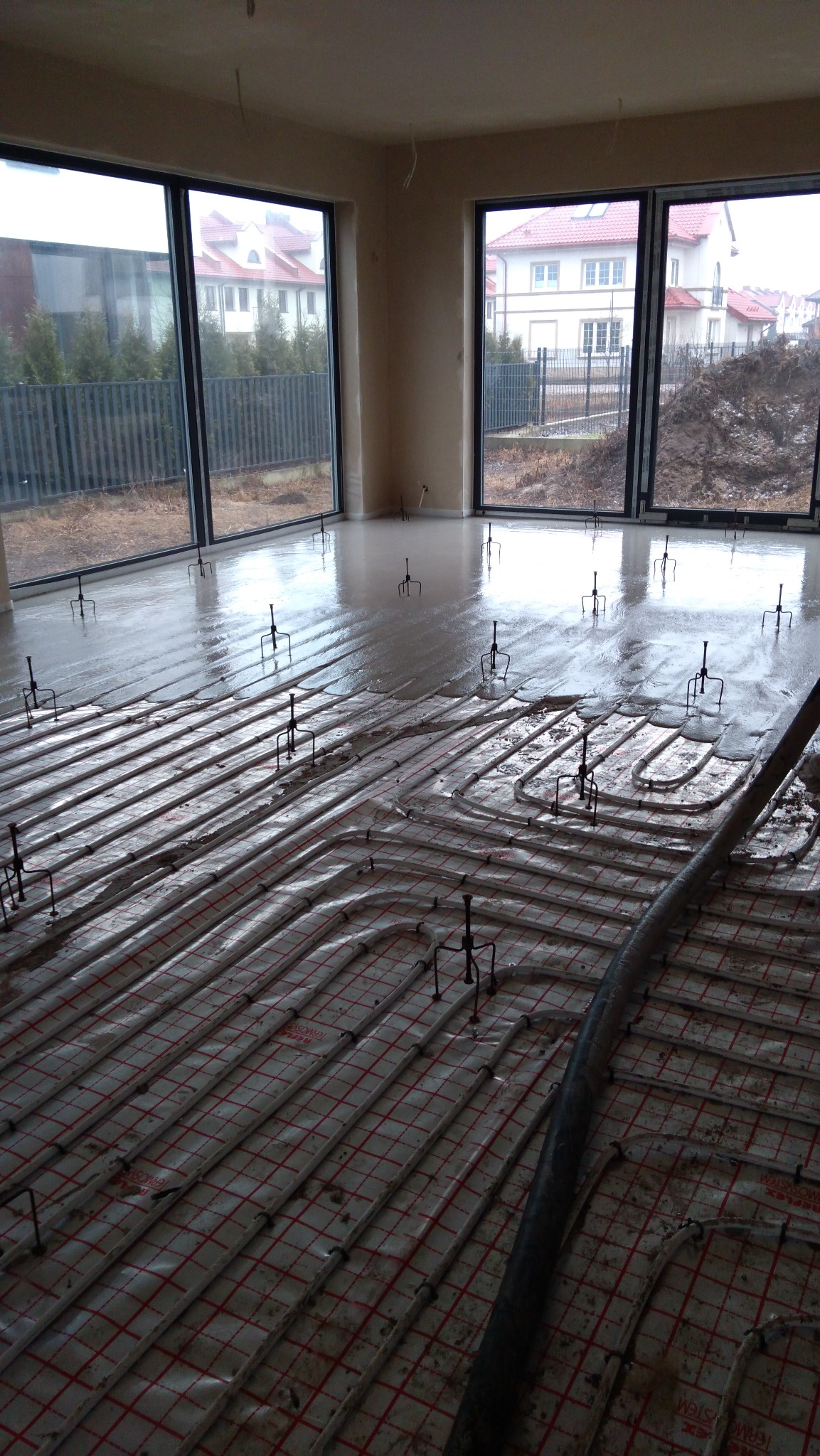 Proces wylewania posadzki w Dębem Wielkim z widocznym systemem ogrzewania podłogowego i elementami poziomującymi, świeżo wylana, mokra powierzchnia odbija światło z dużych okien.