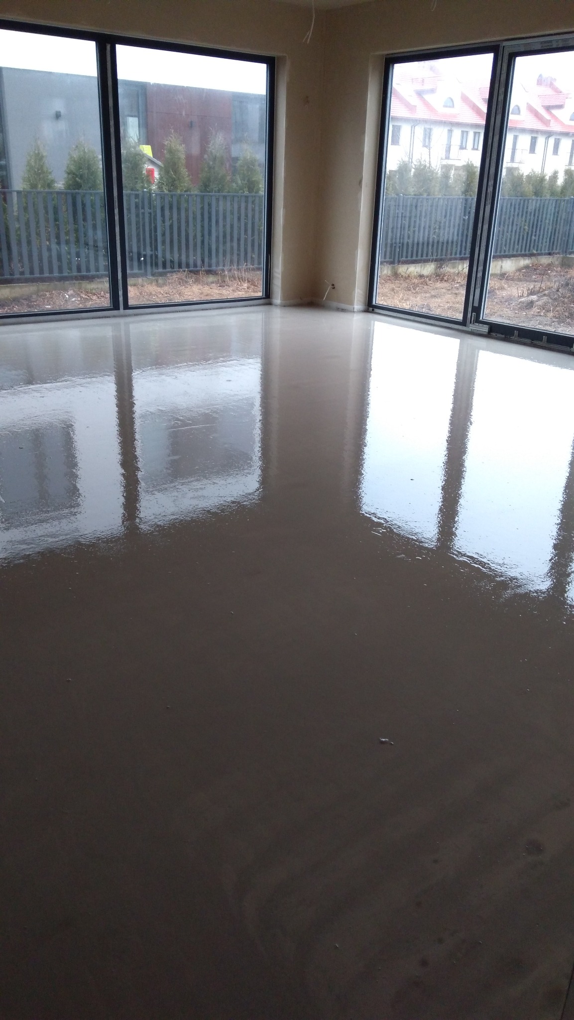 Świeżo wylana, gładka i błyszcząca posadzka jastrychowa w jasnym pomieszczeniu z dużymi oknami, odbijającymi widok zewnętrzny.