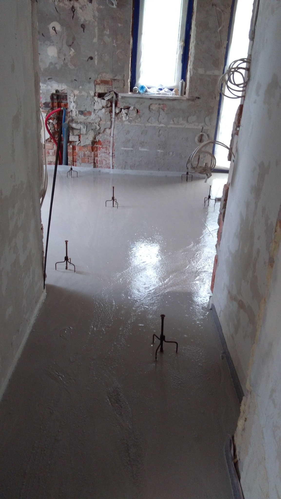 Świeżo wylana, jasnoszara posadzka z widocznymi metalowymi podpórkami poziomującymi, surowe ściany w tle z odsłoniętą cegłą i instalacjami.