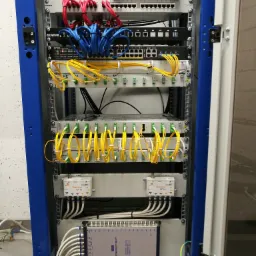 Szafa rack z zainstalowanymi urządzeniami sieciowymi, widoczne patch panele, switche, router, okablowanie światłowodowe i miedziane w kolorach czerwonym, niebieskim i żółtym, oraz multiswitch.
