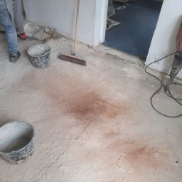 Dominik-KRAK-TRANS-BUD - Wnętrze w trakcie prac remontowych: betonowa podłoga z pyłem, dwa plastikowe wiadra, szczotka do zamiatania i ciężki młot udarowy Hilti TE 1000-AVR przy ścianie.