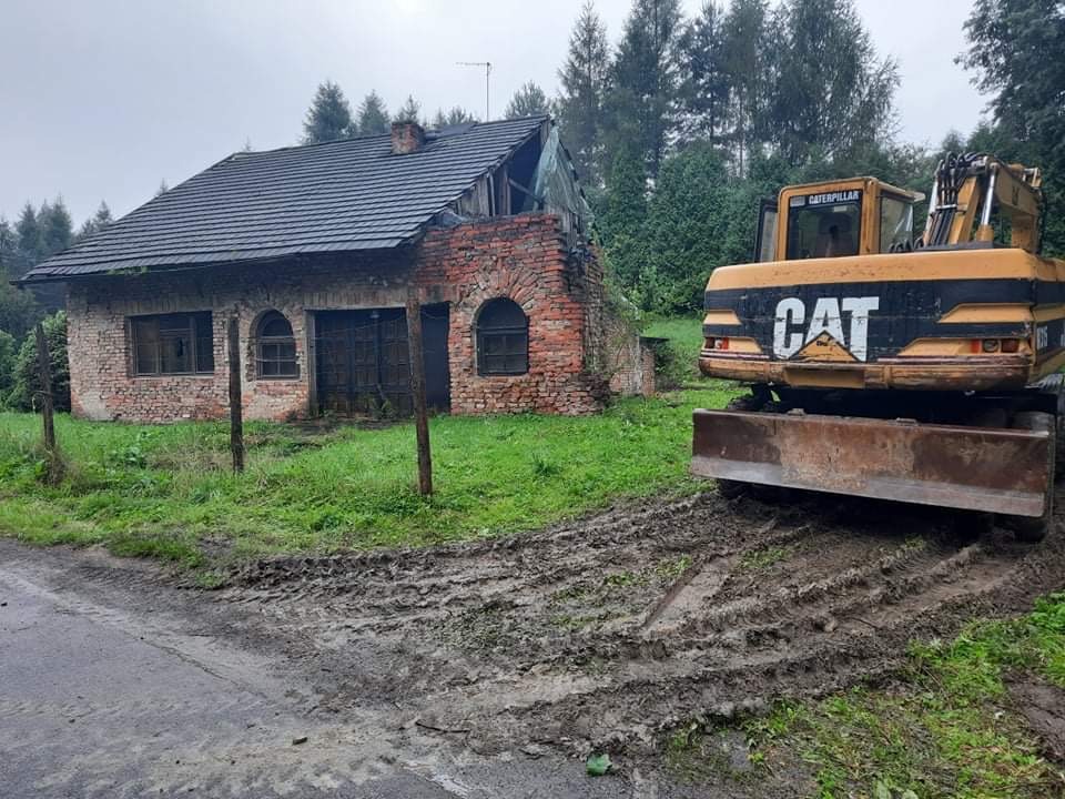 Żółta koparka Caterpillar stoi przed częściowo rozebranym ceglanym budynkiem z ciemnym dachem na zewnątrz w pochmurny dzień, widoczne ślady opon w błocie.