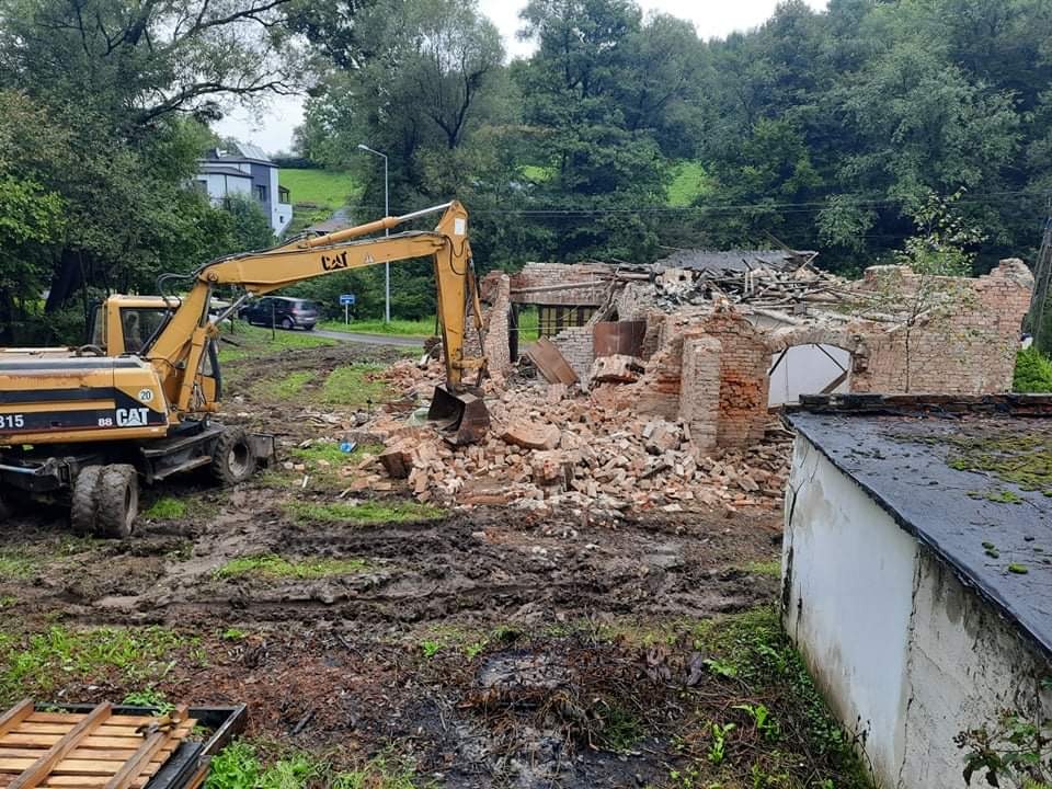 Żółta koparka CAT na błotnistym terenie, gruzowisko po ceglanym budynku w tle, widoczne fragmenty ścian i dachu, drzewa i dom w oddali.