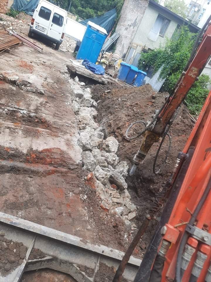 Pomarańczowa koparka z wiertłem pneumatycznym rozbija fundamenty w wykopie; w tle biały bus, niebieska toaleta przenośna i gruz w niebieskich pojemnikach.