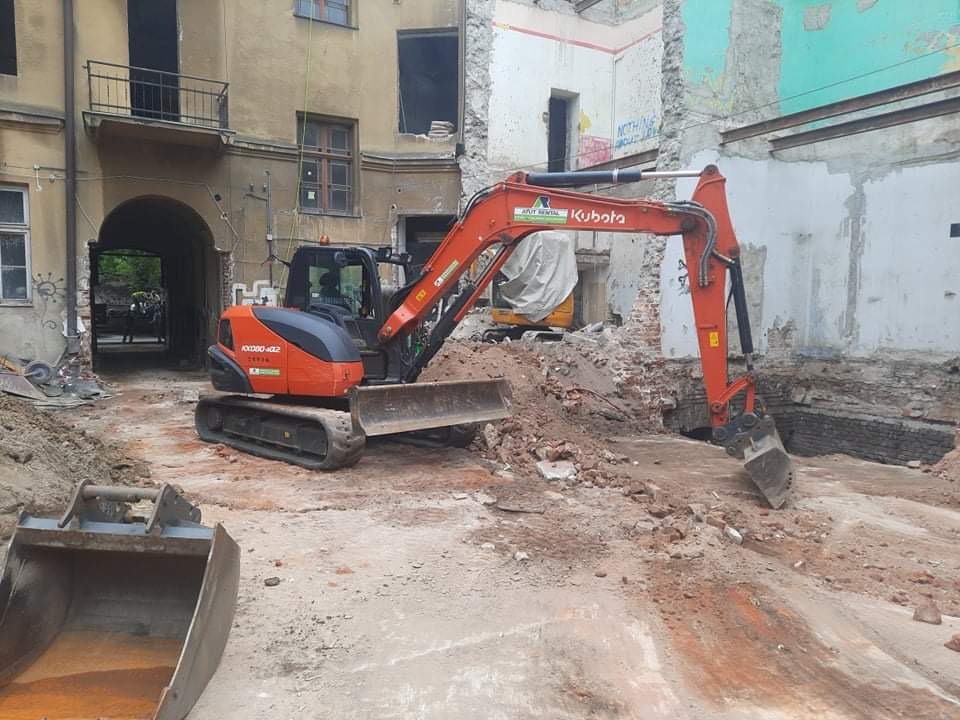 Pomarańczowa minikoparka Kubota przy wykopie na placu budowy w otoczeniu starych budynków z odsłoniętą cegłą i gruzem.