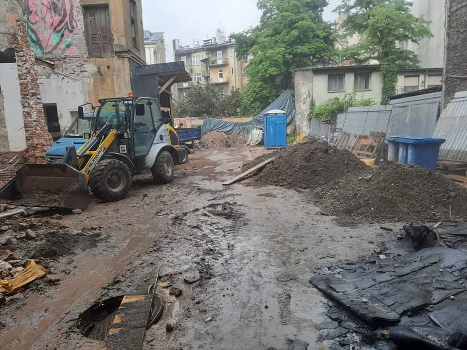 Żółto-czarna ładowarka kołowa na błotnistym placu budowy z gruzem i stertą ziemi, w tle budynek w trakcie rozbiórki i przenośna toaleta.