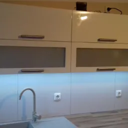 Białe szafki kuchenne na wymiar z podświetleniem LED pod szafkami, widoczny zlew i bateria.