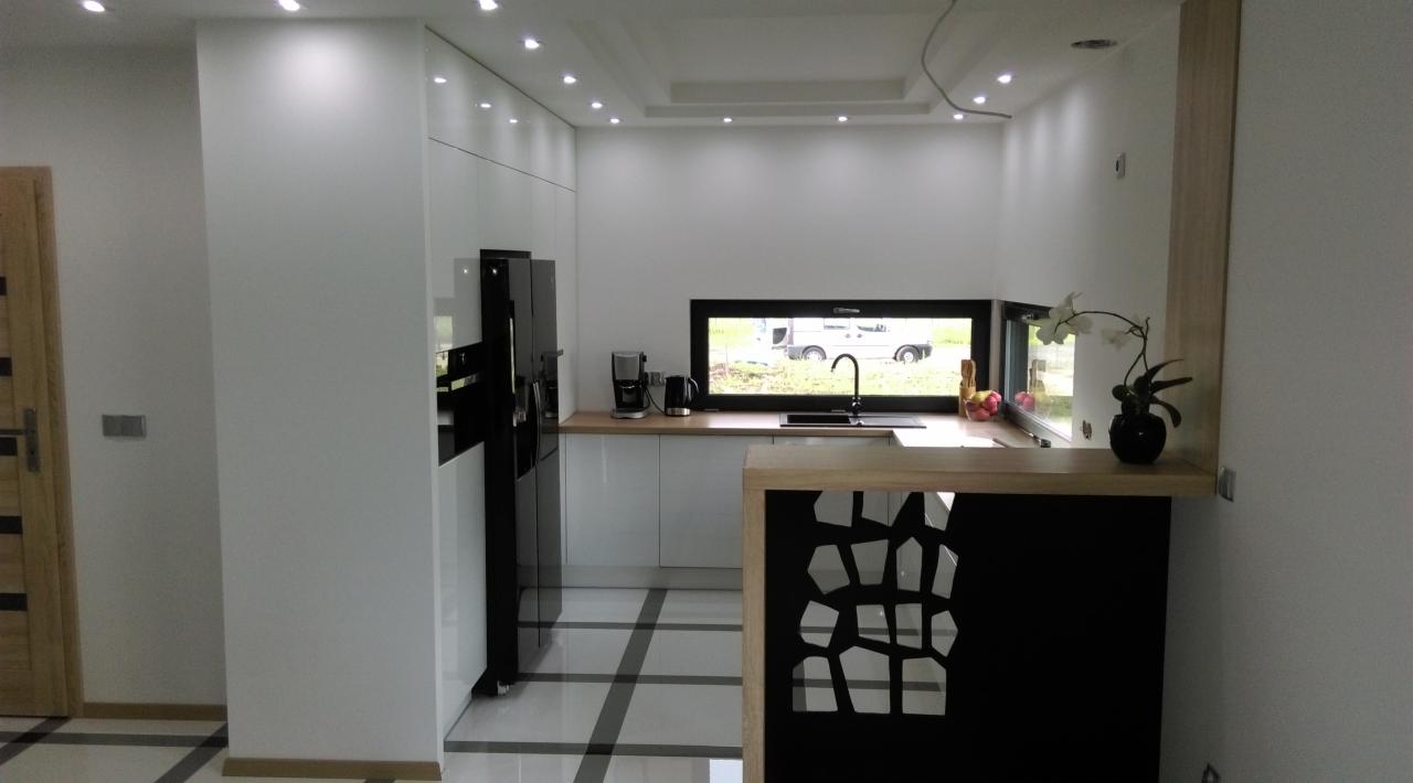 Nowoczesna kuchnia z białymi, błyszczącymi frontami, czarną lodówką i barem z dekoracyjnymi wycięciami, widok z perspektywy wejścia.