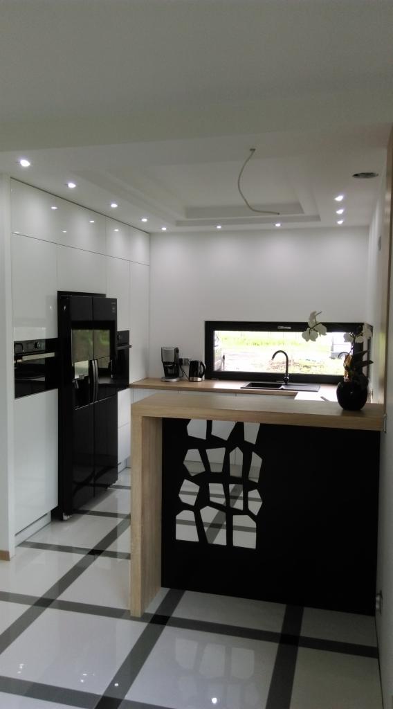 Nowoczesna kuchnia z białymi szafkami, czarną lodówką i wyspą z geometrycznym wzorem na czarnym froncie, wykończona blatem w kolorze jasnego drewna.