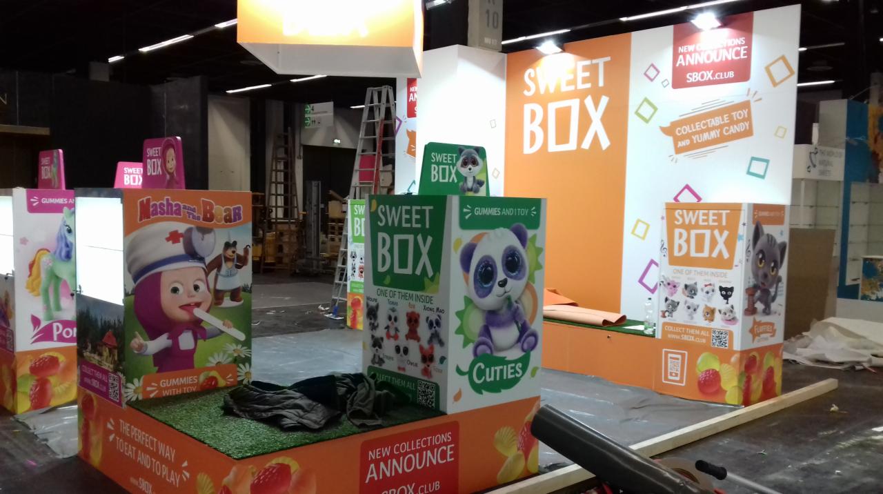 Stoisko targowe z ekspozycją produktów Sweet Box z postaciami z bajek i pluszowymi zabawkami, w trakcie montażu lub demontażu, widoczne elementy konstrukcyjne i narzędzia.