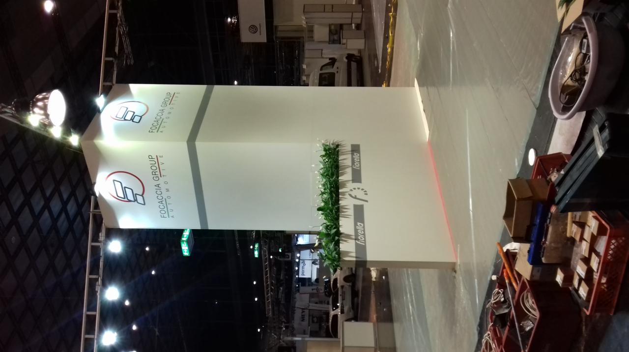 Stoisko targowe z logo Focaccia Group Automotive i dekoracją roślinną Fiorella, widoczne materiały montażowe w skrzynkach.