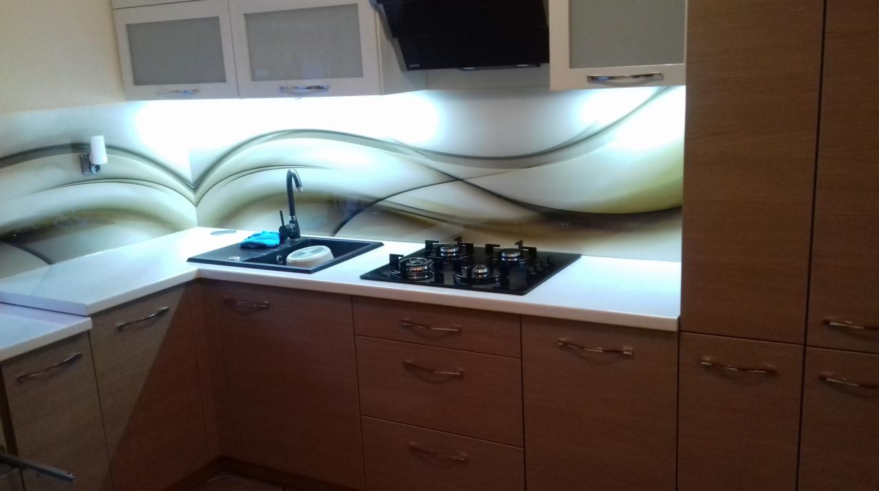 Kuchnia z drewnopodobnymi frontami szafek, białym blatem i czarnym zlewem oraz płytą grzewczą, nad którymi znajduje się panel dekoracyjny z motywem fal.