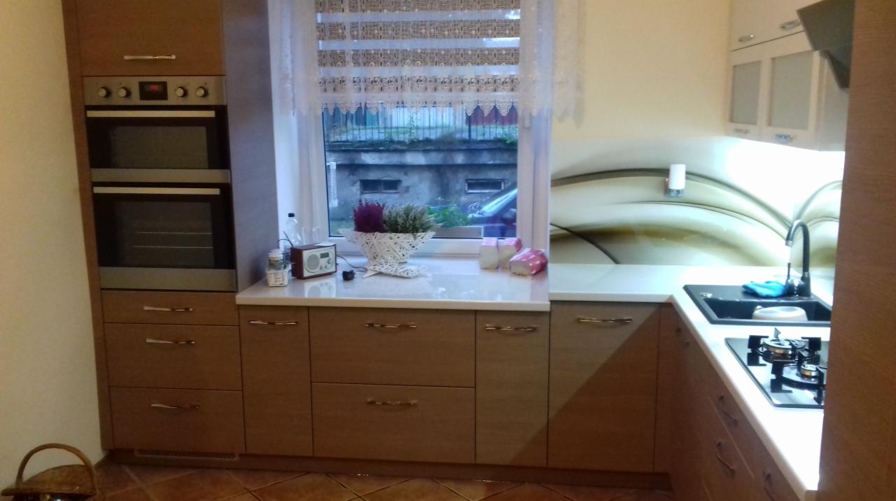 Jasna kuchnia z zabudową meblową: szafki dolne z szufladami, piekarnik w zabudowie, okno z roletą rzymską, czarny zlew i płyta gazowa.