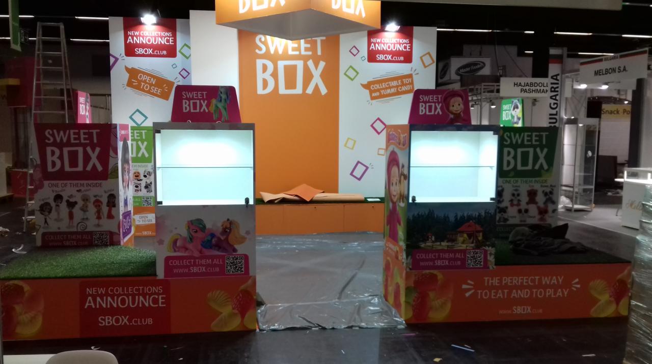 Stoisko targowe marki Sweet Box z podświetlanymi gablotami i grafikami prezentującymi kolekcje zabawek i słodyczy, widoczne materiały reklamowe i drabina w tle.
