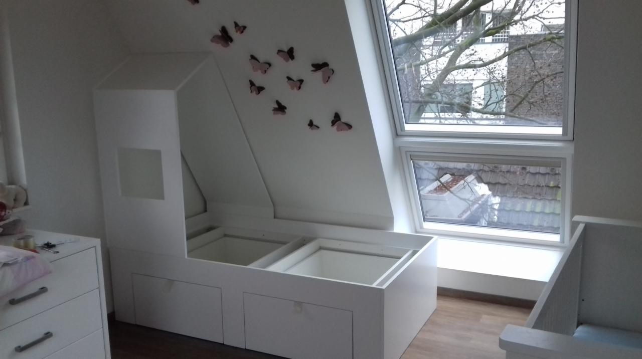 Biała zabudowa pod skosem z szufladami i otwartymi wnękami, motyle dekoracyjne na ścianie, okno dachowe z widokiem na drzewo i budynki.