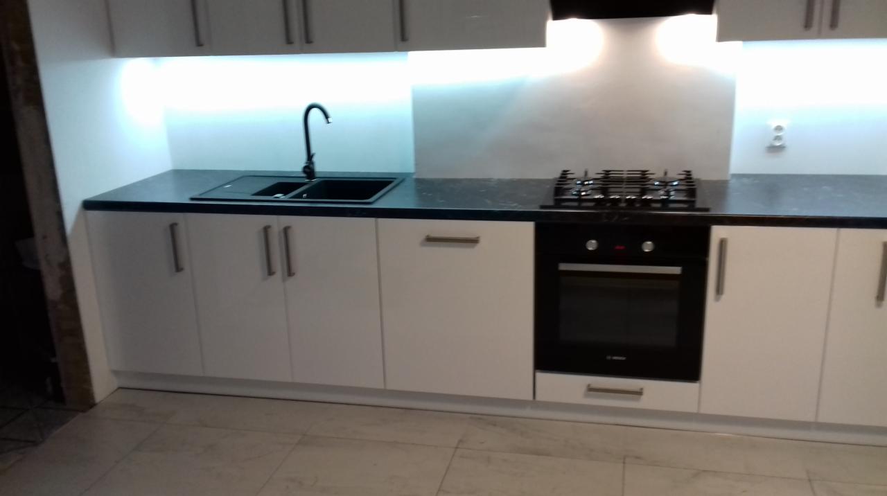 Nowoczesna zabudowa kuchenna z białymi szafkami, czarnym blatem i sprzętem AGD: piekarnik i płyta gazowa, minimalistyczne uchwyty.