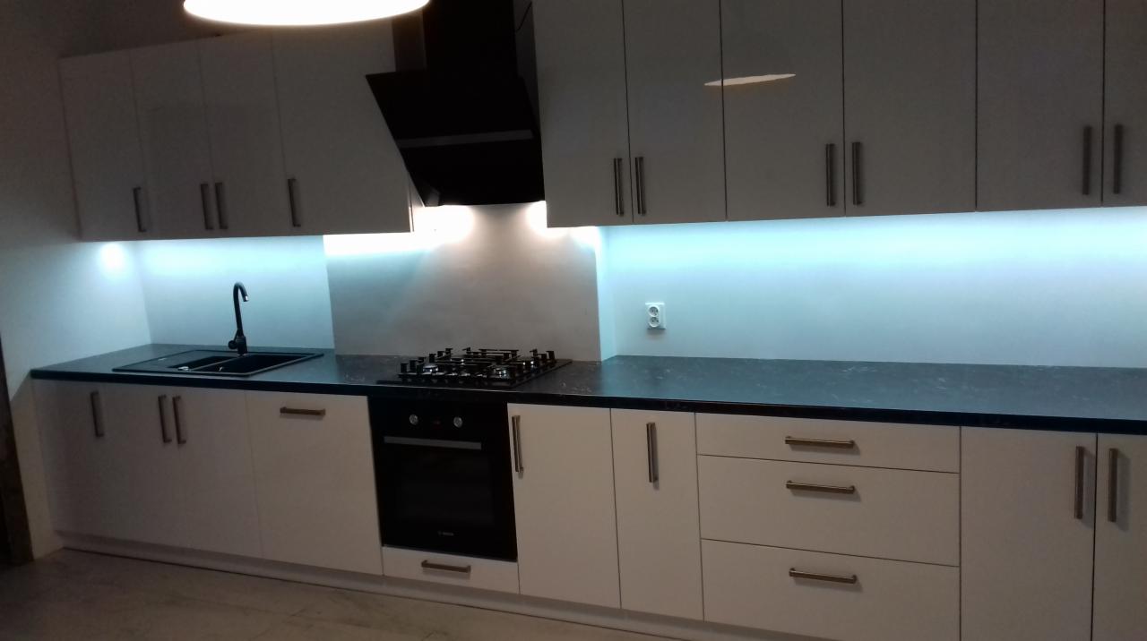 Nowoczesna kuchnia z białymi szafkami, czarnym blatem, płytą grzewczą i piekarnikiem, podświetlona listwą LED pod szafkami wiszącymi.