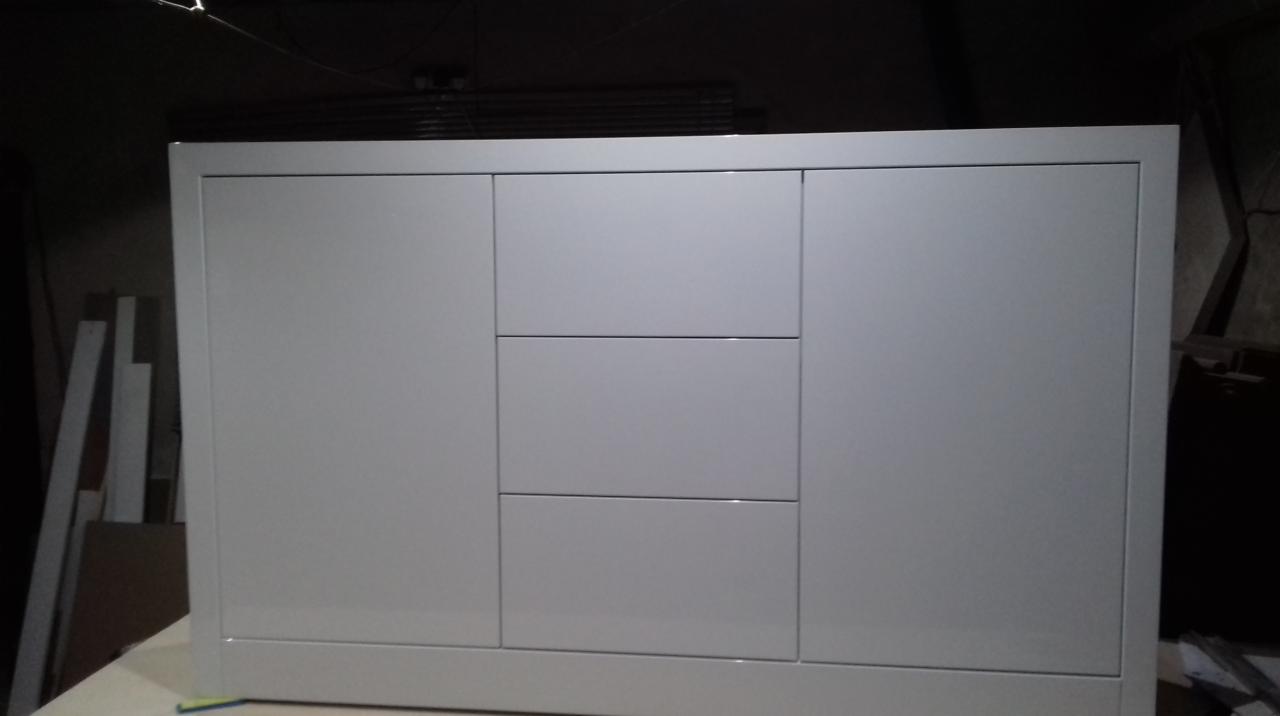 Biała, lakierowana komoda z trzema drzwiami i dwiema szufladami, widoczna w słabym oświetleniu warsztatowym.