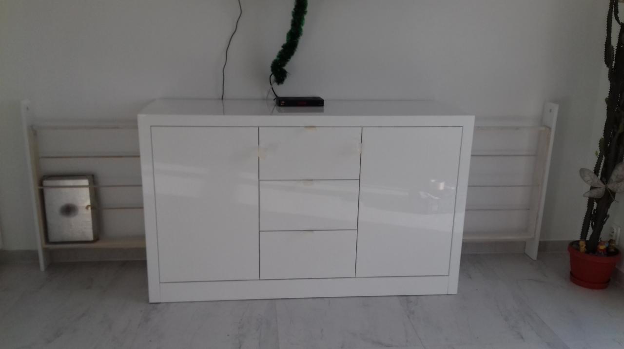 Biała komoda z trzema drzwiami i trzema szufladami, z połyskiem, stojąca na marmurowej podłodze, z widocznym dekoderem na blacie i półkami po bokach.