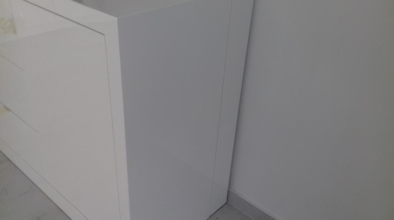 Fragment białej, lakierowanej komody z szufladami, stojącej przy białej ścianie z szarą listwą przypodłogową.