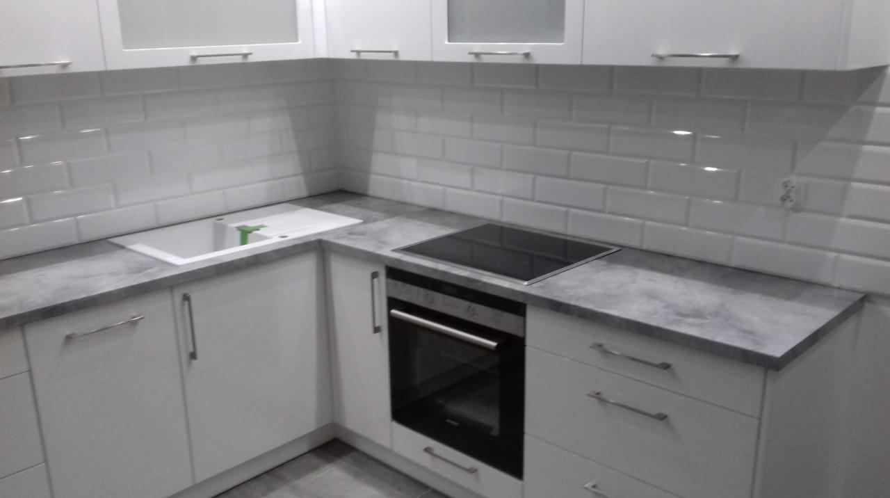 Nowoczesna, biała zabudowa kuchenna z szarym blatem, płytą indukcyjną i piekarnikiem, uzupełniona białą, cegiełkową glazurą.