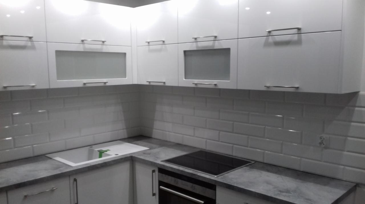 Białe, lakierowane szafki kuchenne z prostokątnymi, metalowymi uchwytami i przeszklonymi frontami, uzupełnione białymi płytkami ściennymi typu metro i szarym blatem.