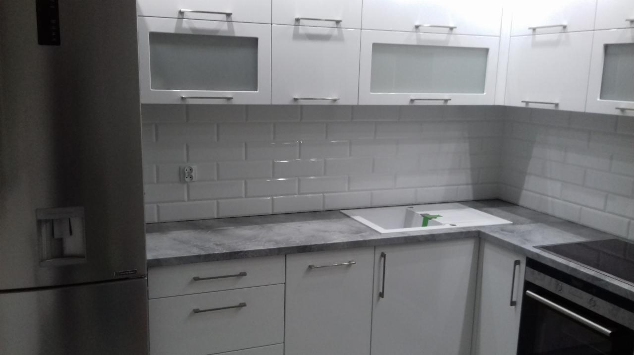 Nowoczesna, biała zabudowa kuchenna z szarym blatem, białymi płytkami typu metro i stalową lodówką po lewej stronie.