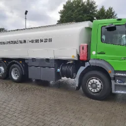 Ciężarówka cysterna w kolorze zielono-białym z napisem 'Hurtowa sprzedaż paliw' oraz numerem telefonu, zaparkowana na mokrej, wybrukowanej powierzchni.