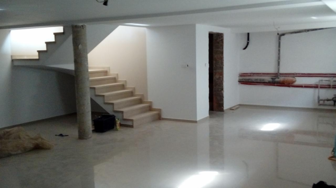 Wnętrze po remoncie: białe ściany, schody z beżowymi stopniami wsparte na okrągłym filarze, błyszcząca podłoga, widoczne rury instalacyjne na ścianie.