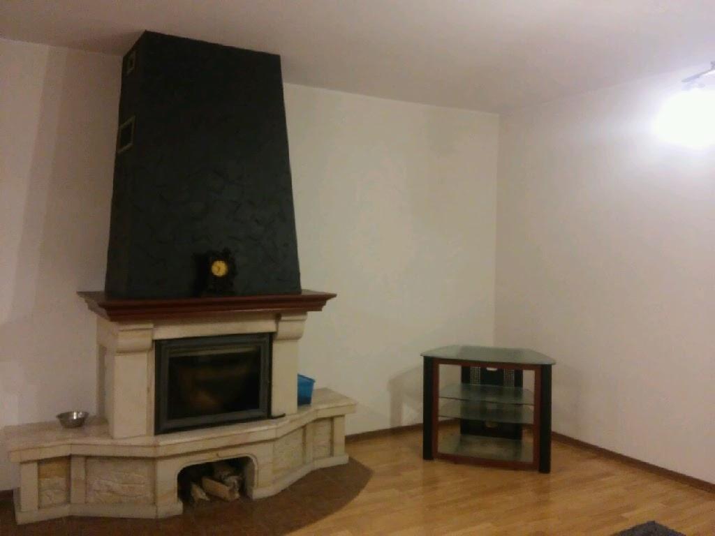 Wykończony kominek z czarnym, teksturowanym nadmurowaniem i jasnym kamiennym obramowaniem, uzupełniony o zegar na kominku, w pokoju z parkietem i ścianami w kolorze złamanej bieli.