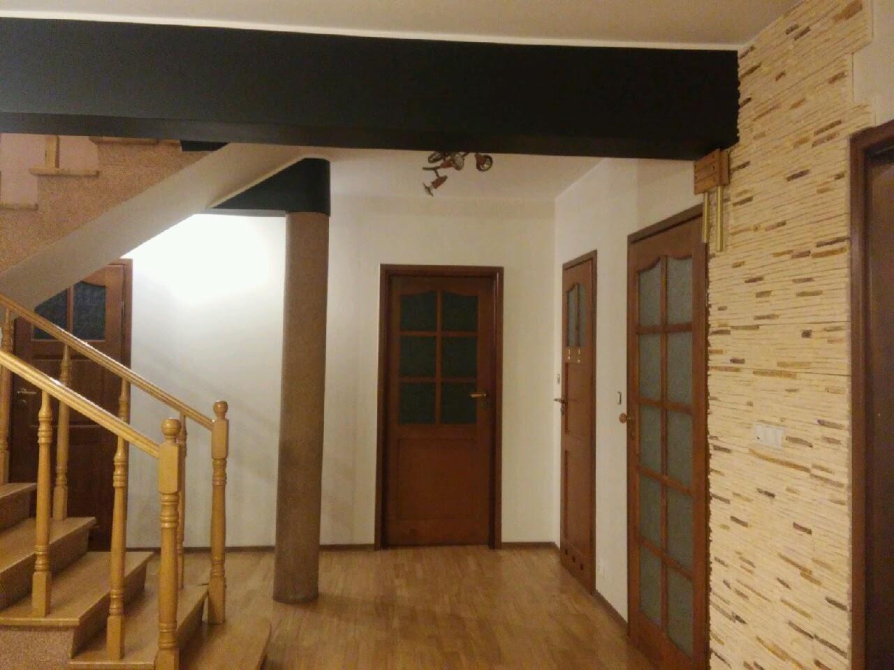 Wnętrze przedpokoju z drewnianymi schodami, trzema drzwiami w różnych odcieniach brązu i ścianą wykończoną dekoracyjnym kamieniem w odcieniach beżu i brązu. Belka stropowa pomalowana na czarno.