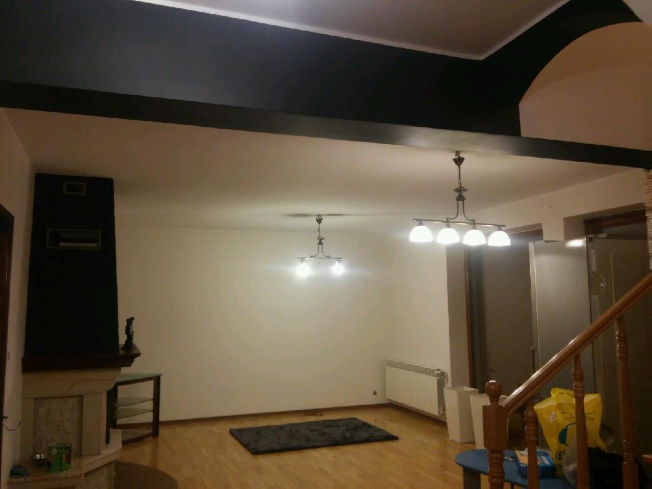 Pomieszczenie po odświeżeniu: białe ściany, czarny sufit i belki, drewniana podłoga, widoczne lampy sufitowe i kominek.