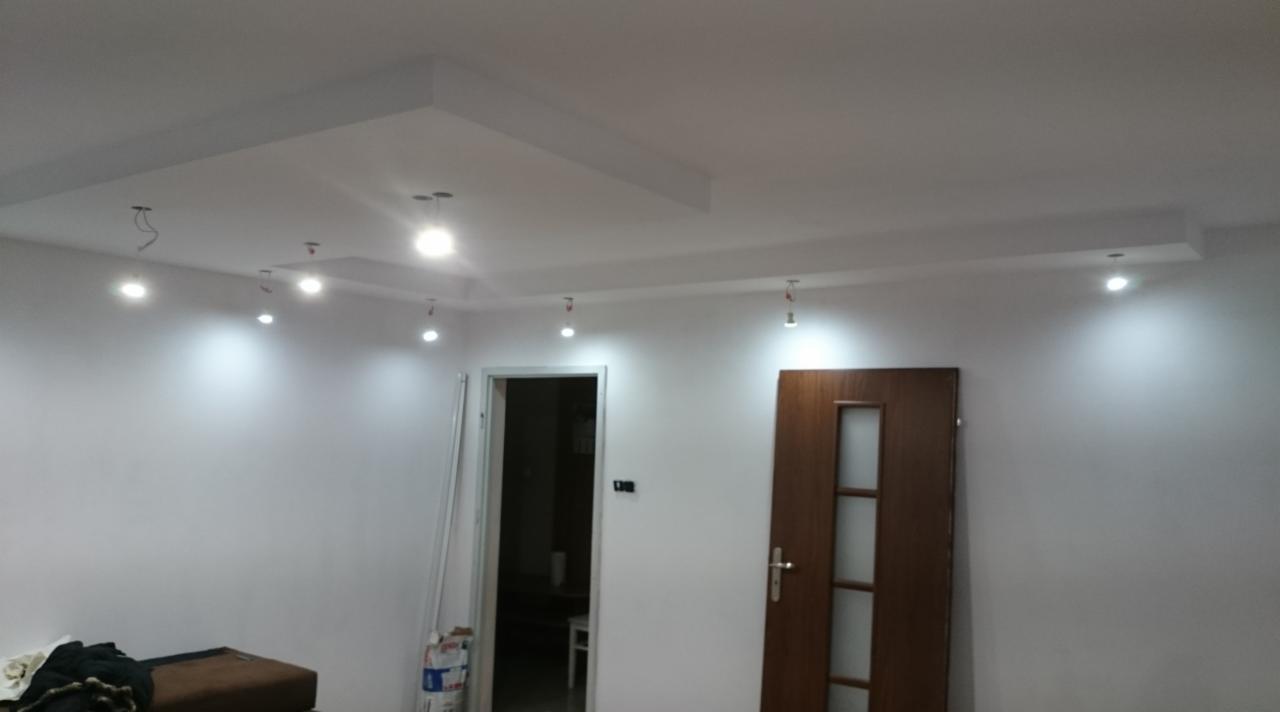 Pokój po odświeżeniu: białe ściany, drewniane drzwi z szybami, sufit z podwieszanym elementem i punktowym oświetleniem, widoczne kable od lamp.