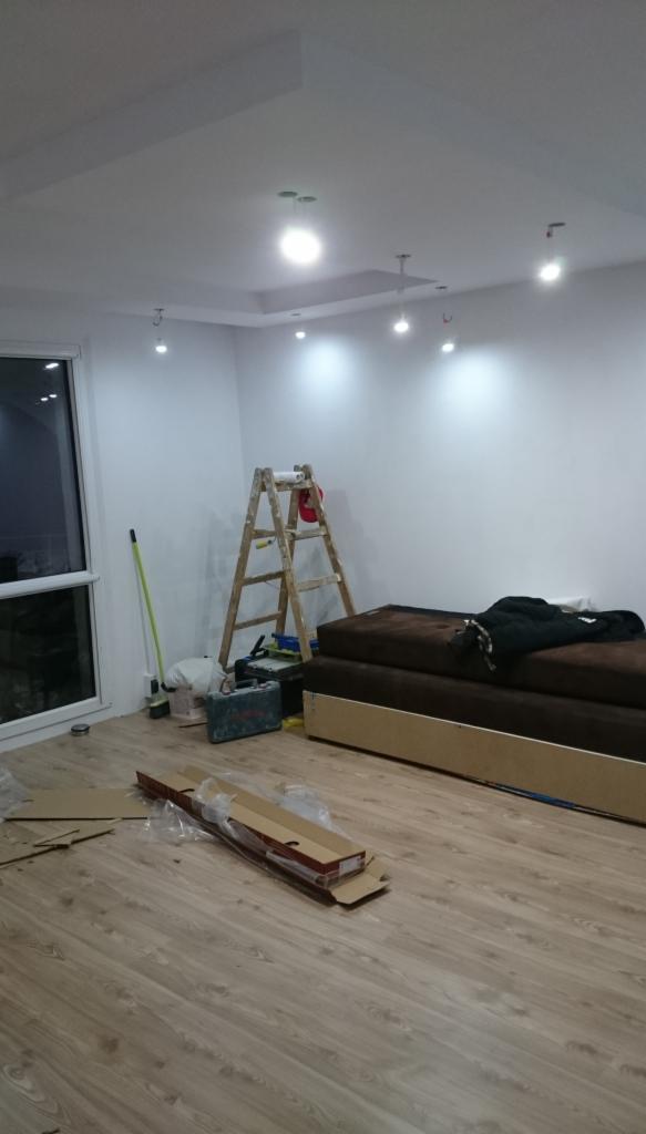 Pokój po remoncie: świeżo pomalowane białe ściany z zamontowanym oświetleniem sufitowym, drabina malarska, narzędzia, podłoga częściowo zakryta kartonami po panelach.