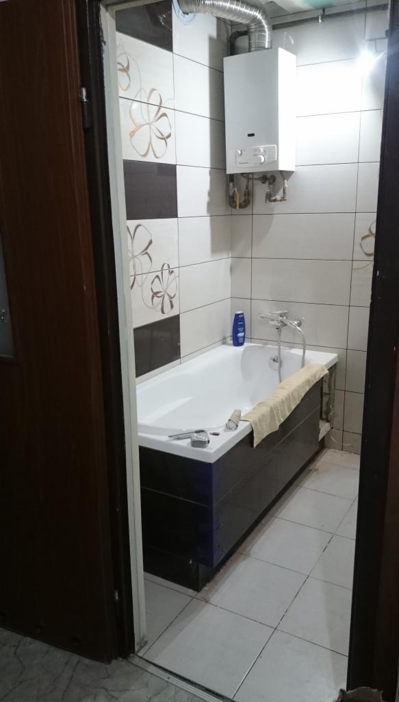 Łazienka po remoncie: biała wanna z ciemną obudową, grzejnik gazowy na ścianie z dekoracyjnymi płytkami, widoczne narzędzia na wannie, ręcznik i butelka płynu do kąpieli.