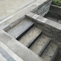 Schody z kostki brukowej z czarnymi obrzeżami, w trakcie zasypywania piaskiem, mur oporowy z kostki po prawej stronie.