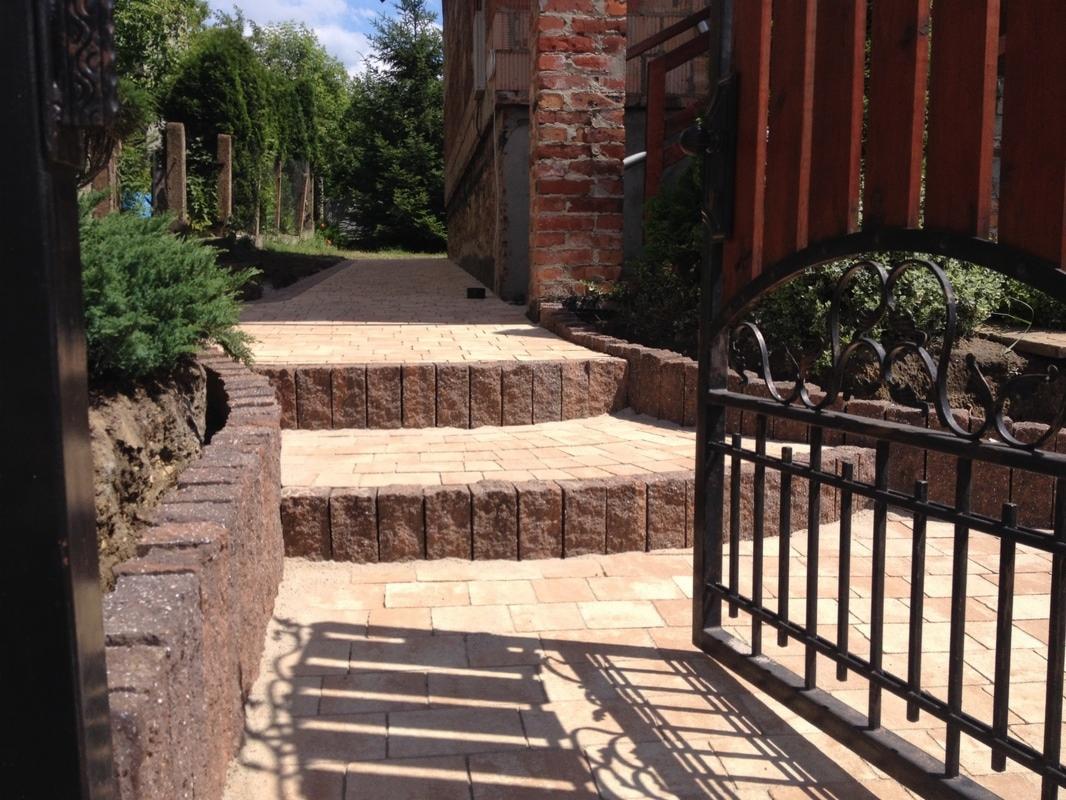 Schody i ścieżka z kostki brukowej prowadzące przez otwartą, ozdobną bramę na posesję z ceglanym budynkiem w tle.