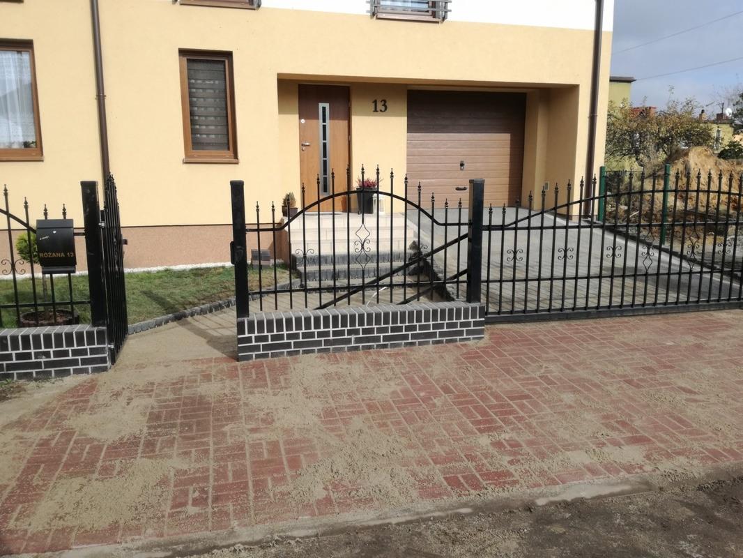 Czerwona kostka brukowa ułożona przed domem z czarnym, kutym ogrodzeniem i bramą, częściowo zasypana piaskiem.