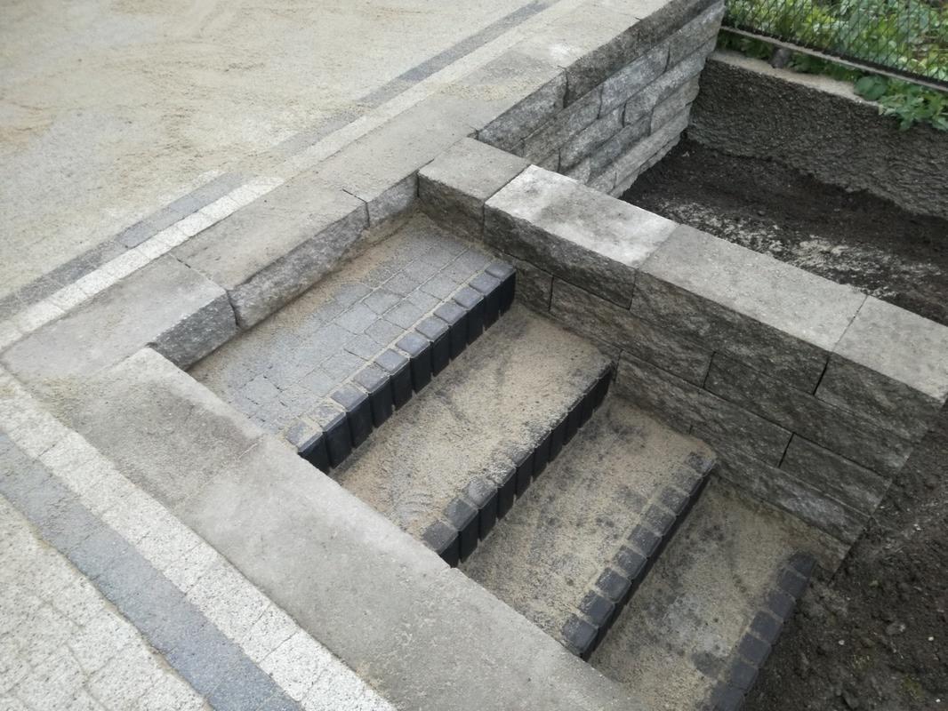 Schody z kostki brukowej z czarnymi obrzeżami, w trakcie zasypywania piaskiem, mur oporowy z kostki po prawej stronie.