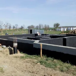 Zabezpieczone hydroizolacją fundamenty domu jednorodzinnego z wystającymi prętami zbrojeniowymi, widoczne elementy szalunków i przygotowane przepusty instalacyjne na tle krajobrazu podmiejskiego.