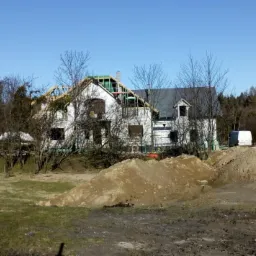 Dom murowany w trakcie budowy z widoczną konstrukcją dachu, ocieplony styropianem, z hałdami ziemi na pierwszym planie i zaparkowanym busem oraz przenośną toaletą w tle.