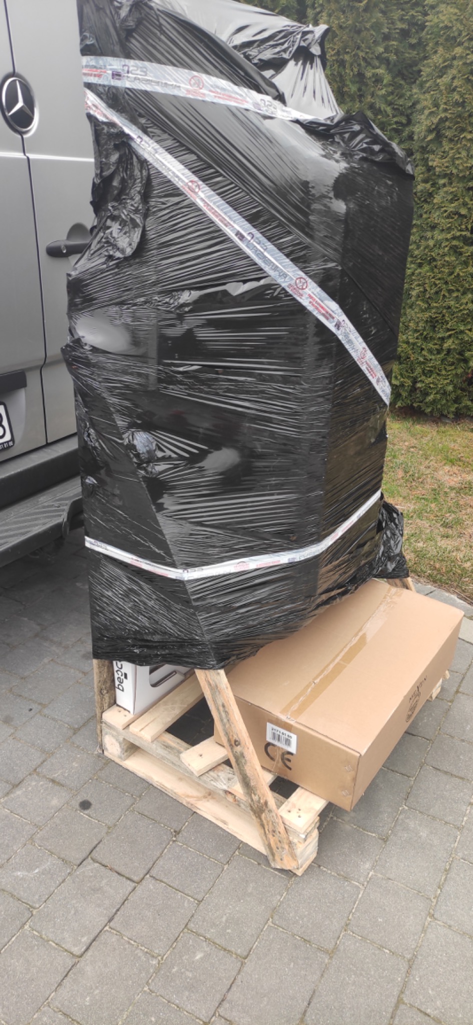Paczka owinięta czarną folią i zabezpieczona taśmą, umieszczona na palecie obok srebrnego samochodu dostawczego Mercedes.