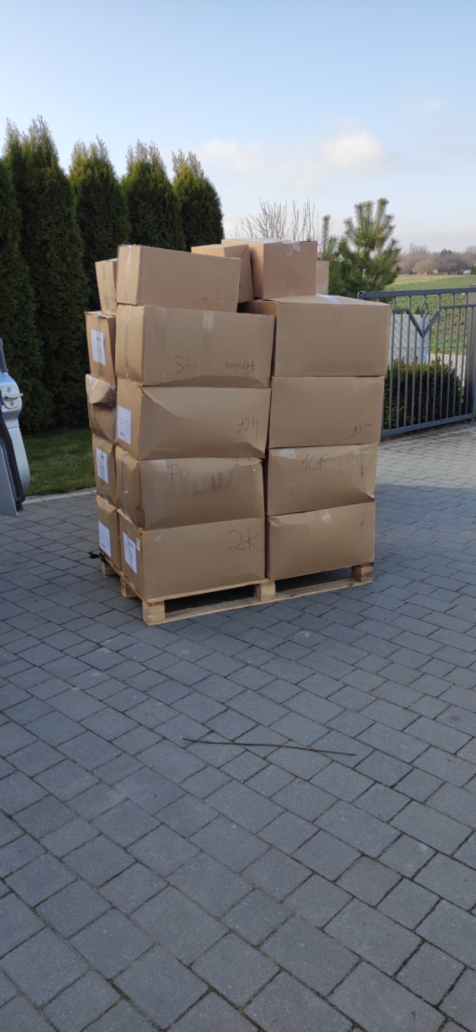 Stos kartonowych pudeł z odręcznymi oznaczeniami, umieszczonych na palecie przed posesją w Zamościu, prawdopodobnie przygotowanych do transportu.