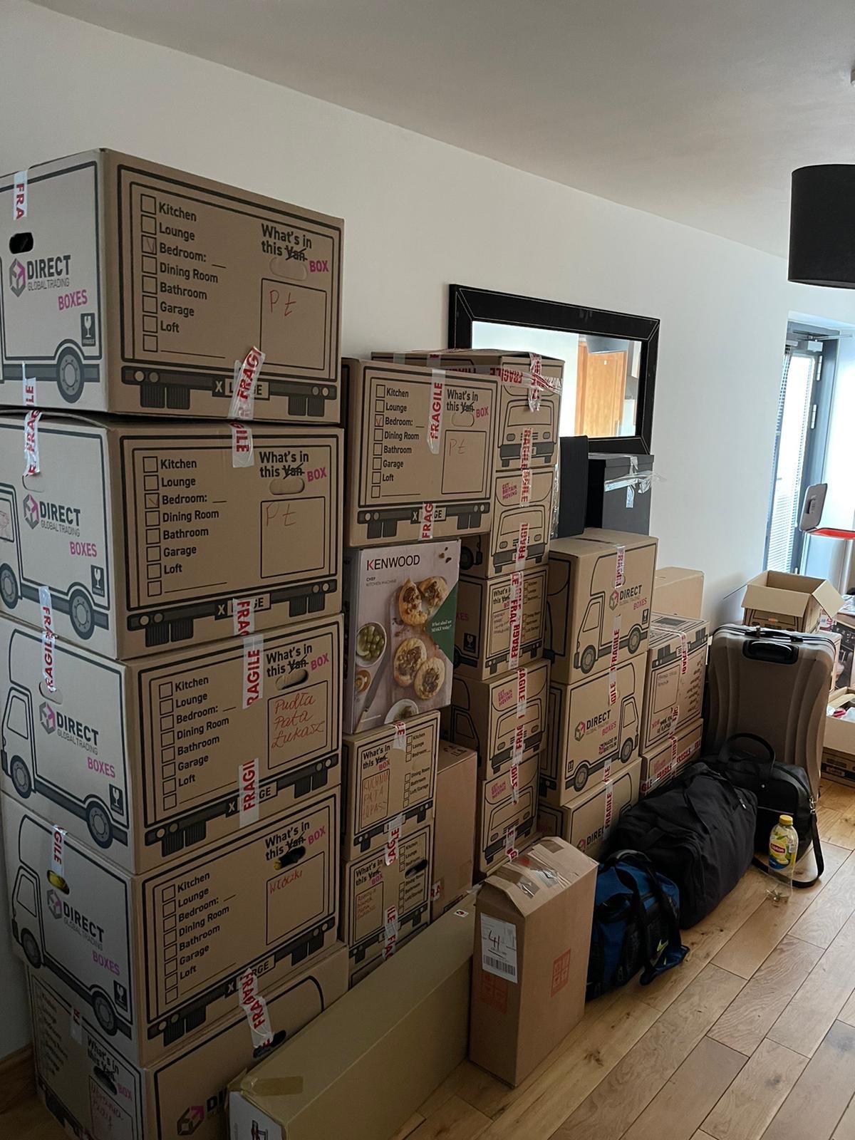 Stos kartonowych pudeł z napisem 'DIRECT GLOBAL TRADING BOXES' i listą pomieszczeń (kuchnia, salon, sypialnia, jadalnia, łazienka, garaż, strych) oraz polem do wypełnienia 'What's in this Van BOX',...