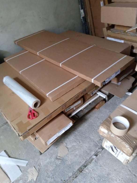 Stos kartonowych pudeł zabezpieczonych białymi paskami, folia stretch, nożyczki i rolka taśmy na podłodze w magazynie, przygotowane do transportu.